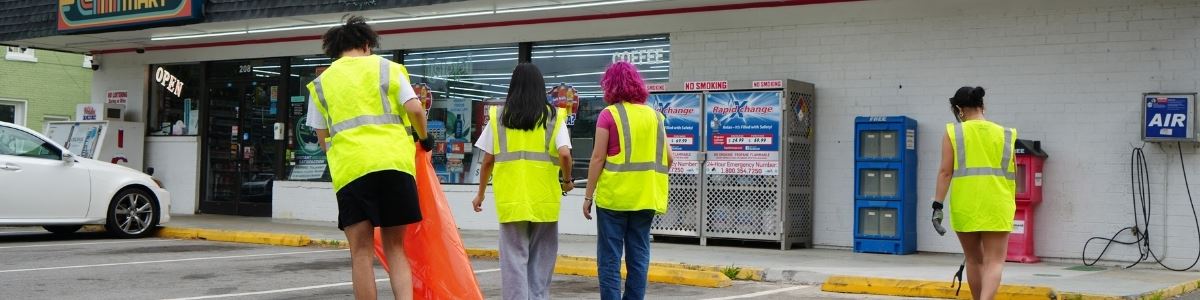 4 teens in neon vests with a neon orange trash bag pick up trash on main street